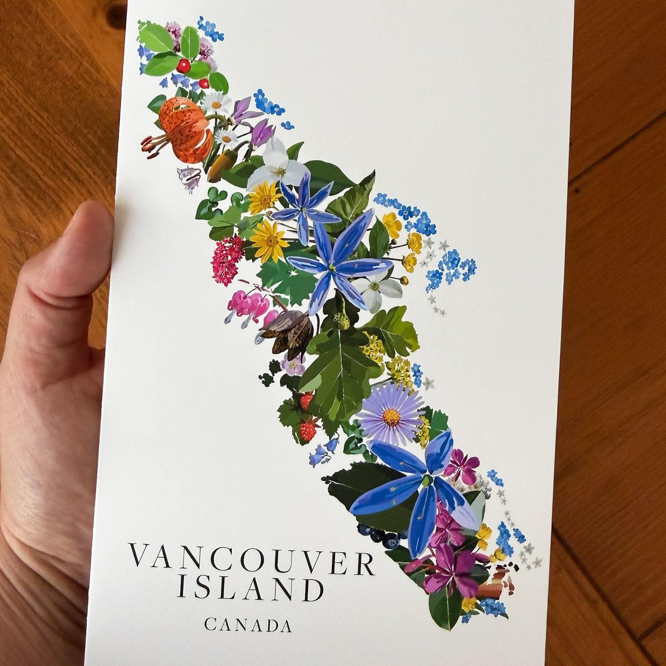 A hand holds a white botanical art greeting card depicting the map of Vancouver Island filled with colorful wildflowers and green foliage, with the text 'VANCOUVER ISLAND CANADA' at the bottom, set against a wooden floor background.