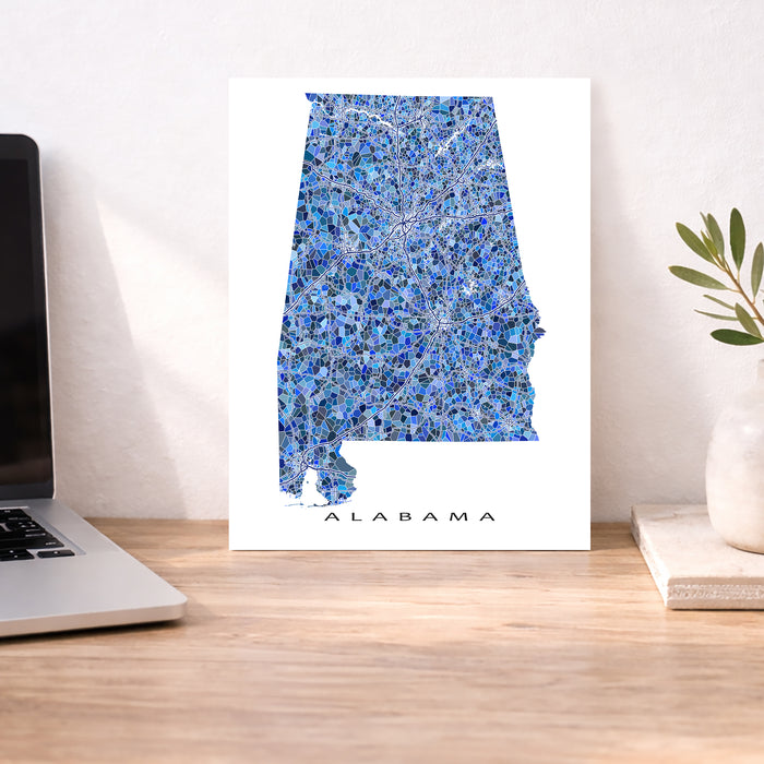 Map of Alabama on a white background next to a laptop and plant on a wooden desk.