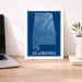 Framed map of Alabama on a desk with a laptop and plant.
