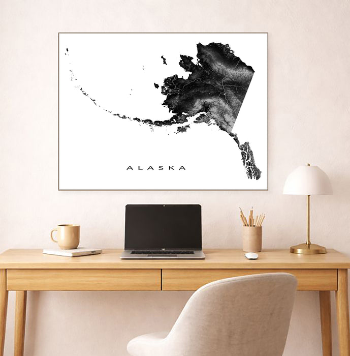 Framed black and white map of Alaska above a desk with a laptop and lamp.