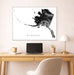 Framed black and white map of Alaska above a desk with a laptop and lamp.