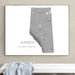 A framed gray topographic map print of Alberta, Canada, displayed on a beige wall above a white bench with a light blue draped cloth.