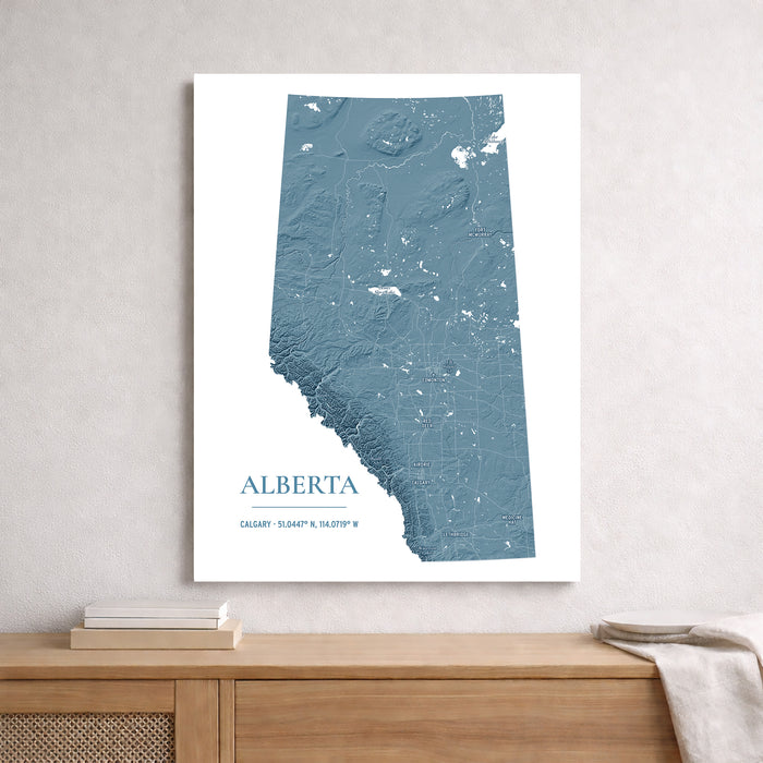 A vertical wall art print featuring a blue-gray relief map of Alberta, Canada, styled on a light gray textured wall above a wooden console table with books and a throw blanket.