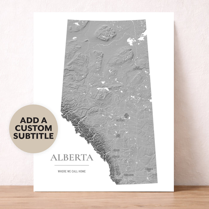 A vertical white poster print featuring a gray topographic map of Alberta, Canada, with the word 'ALBERTA' and smaller text below. The print is leaning against a light beige wall on a wooden surface.
