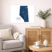A framed Alberta map art print hangs on a beige wall above a light wood cabinet in a styled living room with a gray armchair, books, a mug, and a vase of greenery.