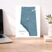 A vertical map print of Alberta, Canada, featuring a textured teal-blue terrain design with white city highlights and the text 'ALBERTA' and 'BORN & raised' at the bottom. The print is displayed on a wooden desk next to a laptop on the left and a small potted plant in a white vase on the right, set against a neutral light beige wall.