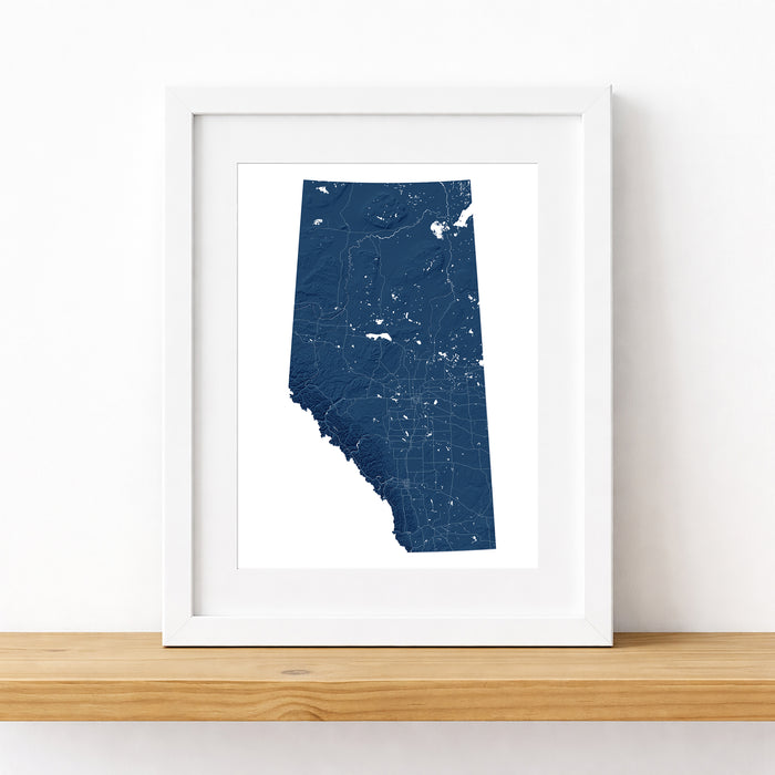 A framed map print of the province of Alberta in dark blue, displayed in a white frame on a light wood shelf against a white wall.