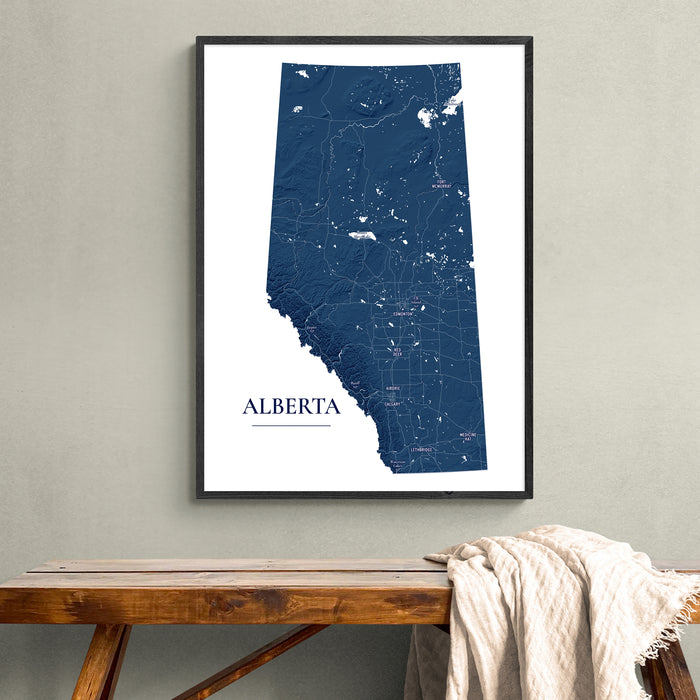 A framed vertical map print of Alberta, Canada, featuring a dark navy blue state silhouette with white road details and the word 'ALBERTA' at the bottom, displayed on a light gray wall above a wooden bench with a draped cream throw blanket.