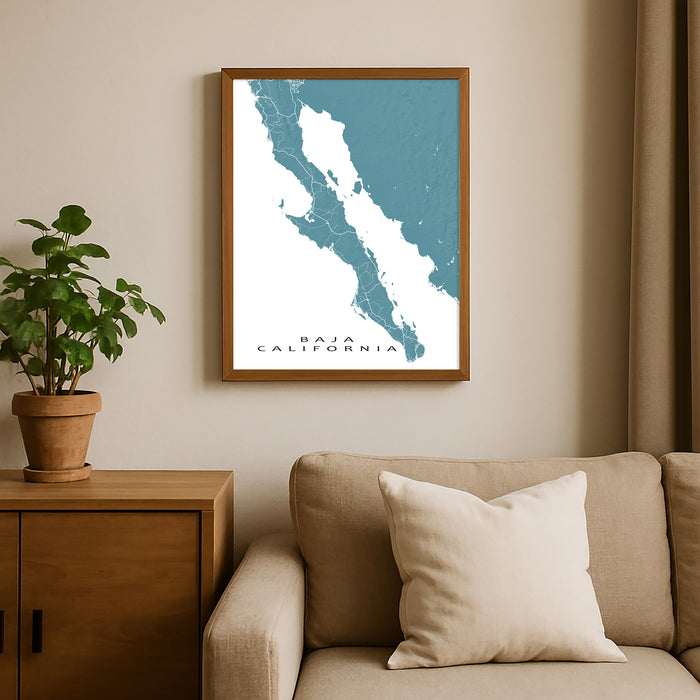 Framed map of Baja California on a wall above a beige sofa with a plant beside it.