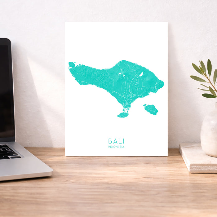 Teal map of Bali on a white poster on a desk with a laptop and plant.
