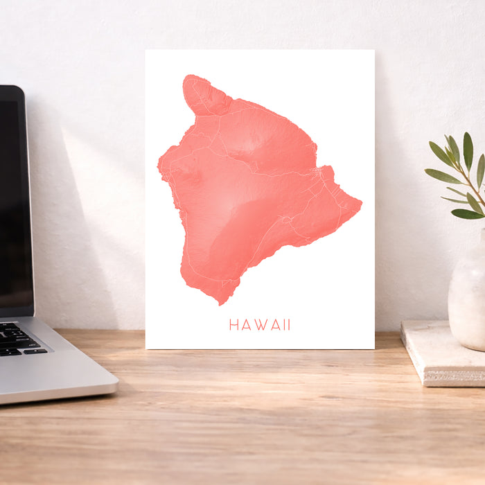Map of Hawaii in coral color on a white background, displayed on a wooden desk with a laptop and plant.