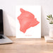 Map of Hawaii in coral color on a white background, displayed on a wooden desk with a laptop and plant.