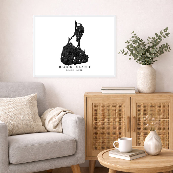 A framed black and white art print of Block Island, Rhode Island, displayed on a light beige wall in a styled living room setting with a gray armchair, wooden cabinet, and greenery.