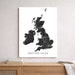 A vertical wall art print showing a black line map of the British Isles on a white background, hanging on a light gray wall above a wooden console table with books and a throw blanket.