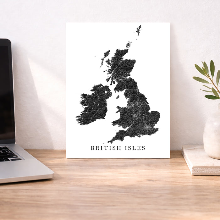 A minimalist black-and-white map print of the British Isles with the text 'BRITISH ISLES' at the bottom, displayed on a wooden desk between a laptop and a small plant in a vase.