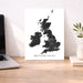 A minimalist black-and-white map print of the British Isles with the text 'BRITISH ISLES' at the bottom, displayed on a wooden desk between a laptop and a small plant in a vase.