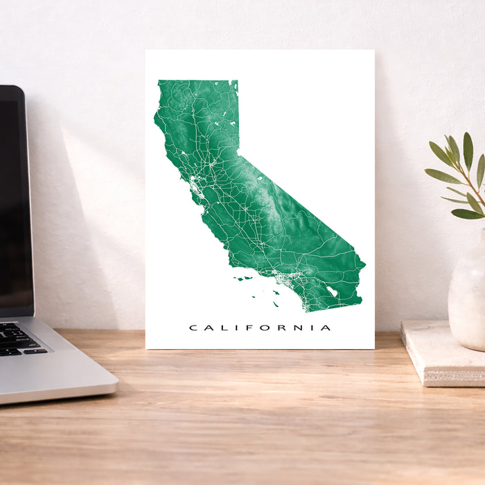 Map of California on a white background displayed on a wooden desk with a laptop and plant.