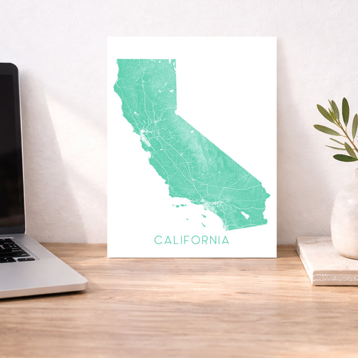 Teal map of California on a white background, displayed on a wooden desk with a laptop and plant.