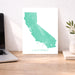 Teal map of California on a white background, displayed on a wooden desk with a laptop and plant.