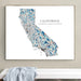 A framed art print of a stylized California map with Los Angeles coordinates, hanging on a beige wall above a white bench with a light blue cloth.