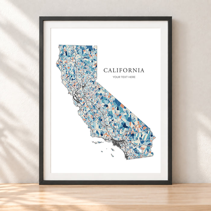 A framed California state map art print with a black frame, leaning against a light beige wall on a wooden surface. The map features a mosaic pattern in shades of blue, teal, and orange on a white background, with the text 'CALIFORNIA' and 'YOUR TEXT HERE'.