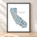 A framed California state map art print with a black frame, leaning against a light beige wall on a wooden surface. The map features a mosaic pattern in shades of blue, teal, and orange on a white background, with the text 'CALIFORNIA' and 'YOUR TEXT HERE'.