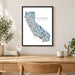 A framed California map art print hangs on a light beige wall above a wooden desk with a mug, pencil cup, books, and plants in a styled home office setting.