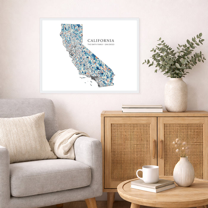 A framed California state map art print hangs on a light beige wall above a gray armchair and a wooden cabinet in a styled living room setting.