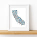 A framed art print of a stylized map of California, featuring a multicolor mosaic pattern in shades of blue, beige, and white. The print is housed in a clean white frame and displayed on a light wood shelf against a white wall.
