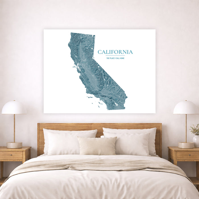 A large framed wall art print featuring a blue-green topographic map of California with the text 'CALIFORNIA' and 'THE PLACE TO GO HOME' displayed above a neatly made bed in a modern bedroom setting.