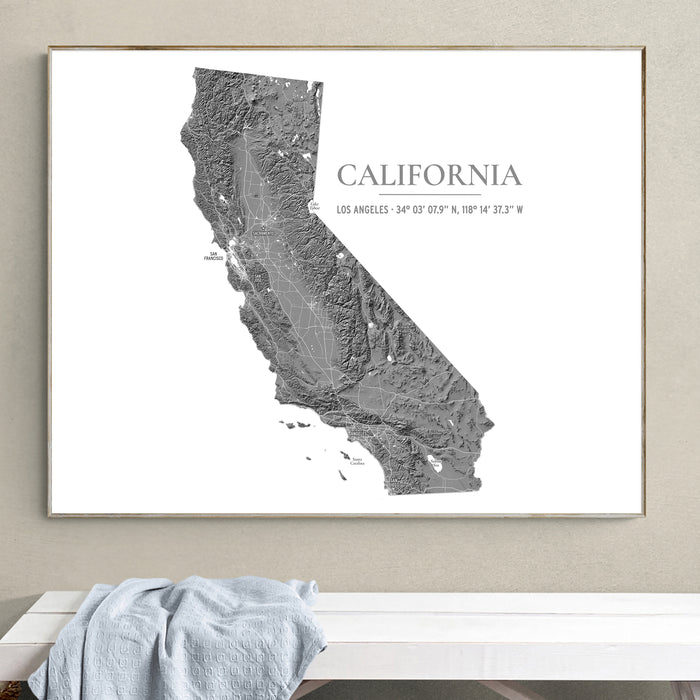 A framed California state map art print in grayscale, hanging on a light beige wall above a white bench with a light blue draped cloth.