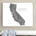 A framed California state map art print in grayscale, hanging on a light beige wall above a white bench with a light blue draped cloth.
