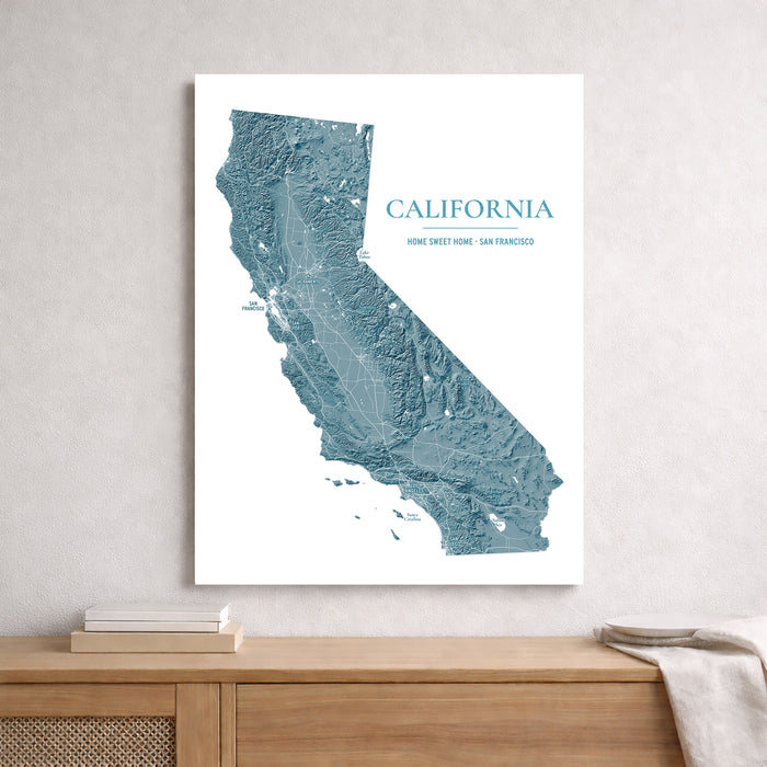 A vertical white wall art print featuring a blue-gray topographic map of California with the text 'CALIFORNIA' and 'HOME SWEET HOME SAN FRANCISCO,' displayed above a wooden console table against a light gray wall.