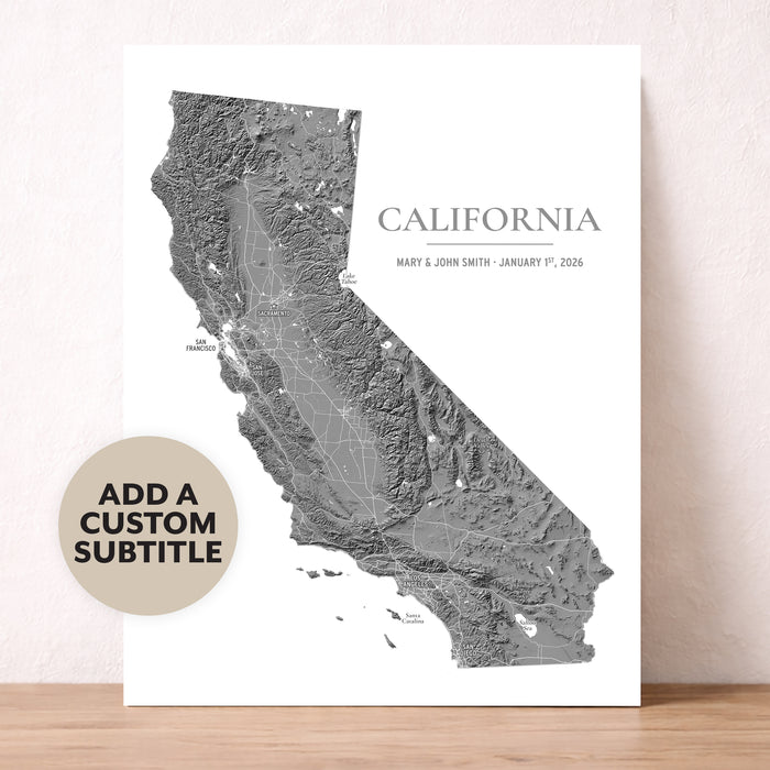 A custom California map art print with a gray topographic design and text, displayed leaning against a textured beige wall on a light wood surface. A circular badge on the left says 'ADD A CUSTOM SUBTITLE'.