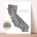 A custom California map art print with a gray topographic design and text, displayed leaning against a textured beige wall on a light wood surface. A circular badge on the left says 'ADD A CUSTOM SUBTITLE'.