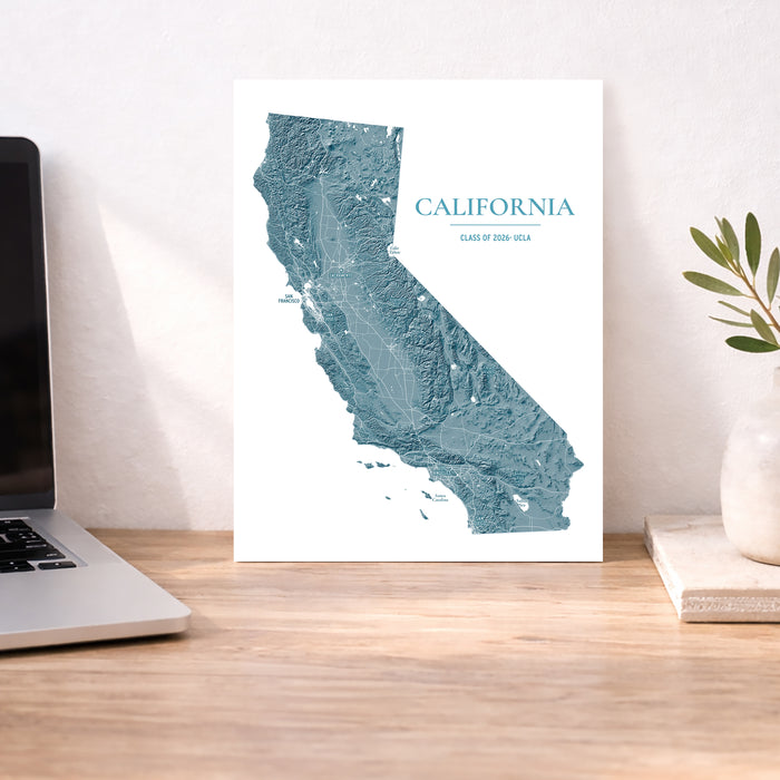 A vertical wall art print featuring a blue-gray relief map of California with the text 'CALIFORNIA' and 'DESERT OF USA' on a white background, displayed on a wooden desk next to a laptop and a small plant.