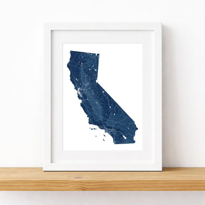 A framed California state map print in dark blue with white linework, displayed in a white frame on a light wood shelf against a white wall.