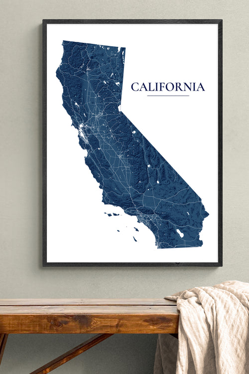 A framed California state map art print in dark blue and white, hanging on a light gray wall above a rustic wooden bench with a draped cream throw blanket.