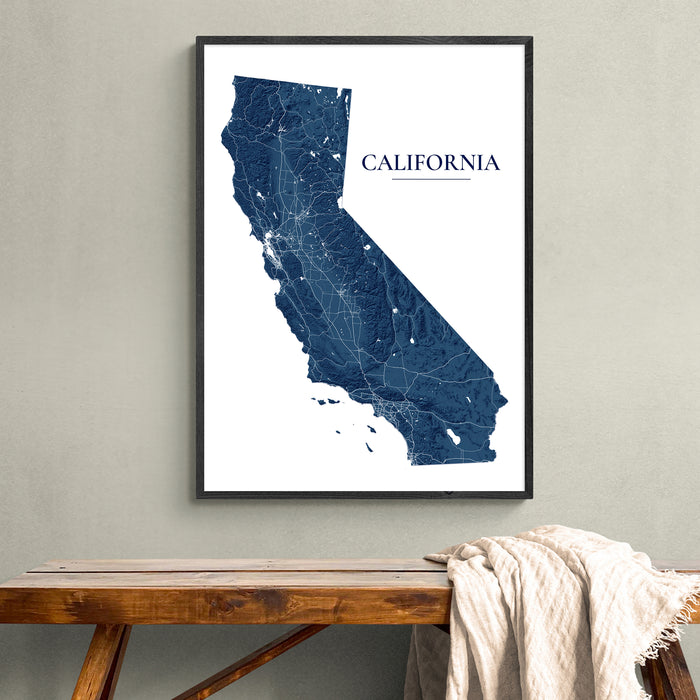 A framed California state map art print in dark blue and white, hanging on a light gray wall above a rustic wooden bench with a draped cream throw blanket.