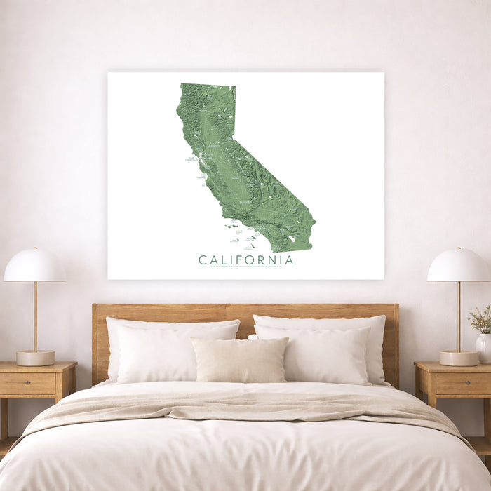 A framed wall art print showing a green topographic map of California with the word CALIFORNIA underneath, displayed above a neatly made bed in a bright bedroom.