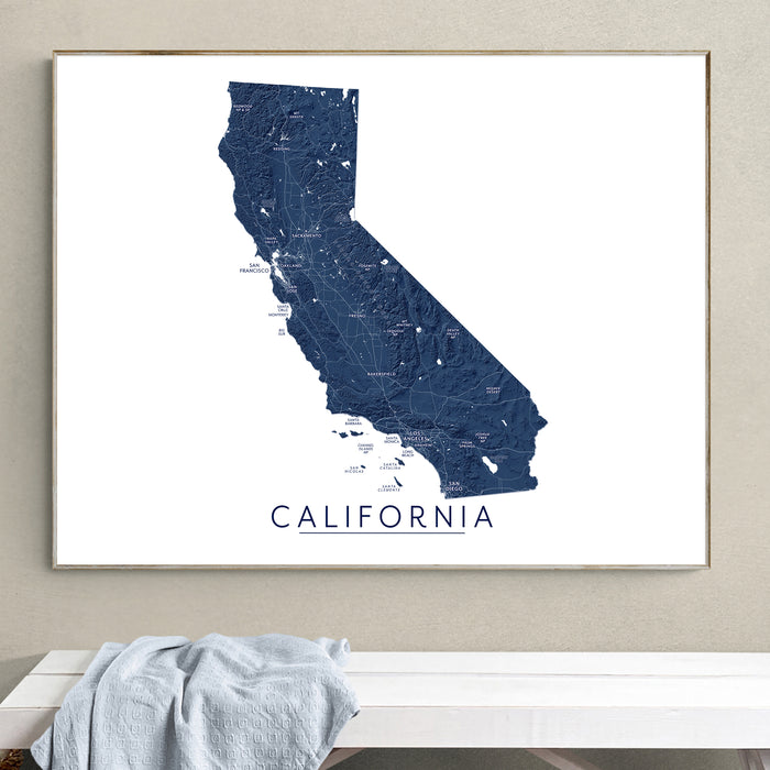 A framed California state map print in dark blue and white, hanging on a beige wall above a white bench with a light blue cloth draped over it.