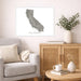 A framed gray map print of California hangs on a light beige wall above a gray armchair and a wooden cabinet in a styled living room.