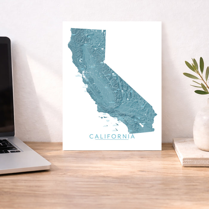 A vertical wall art print featuring a teal-blue topographic map of California with the word 'CALIFORNIA' at the bottom, displayed on a wooden desk beside a laptop and a small plant in a vase.