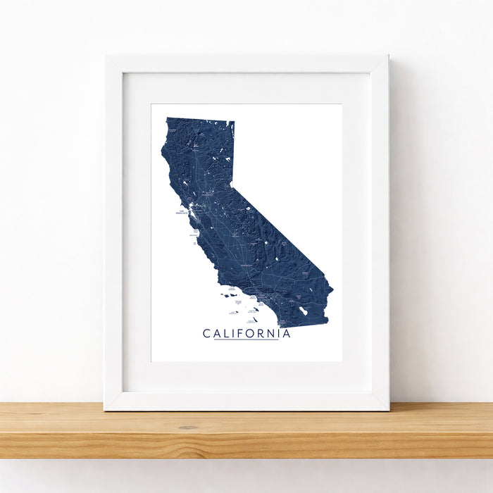 A framed map print of California in dark blue, displayed on a light wood shelf against a white wall.