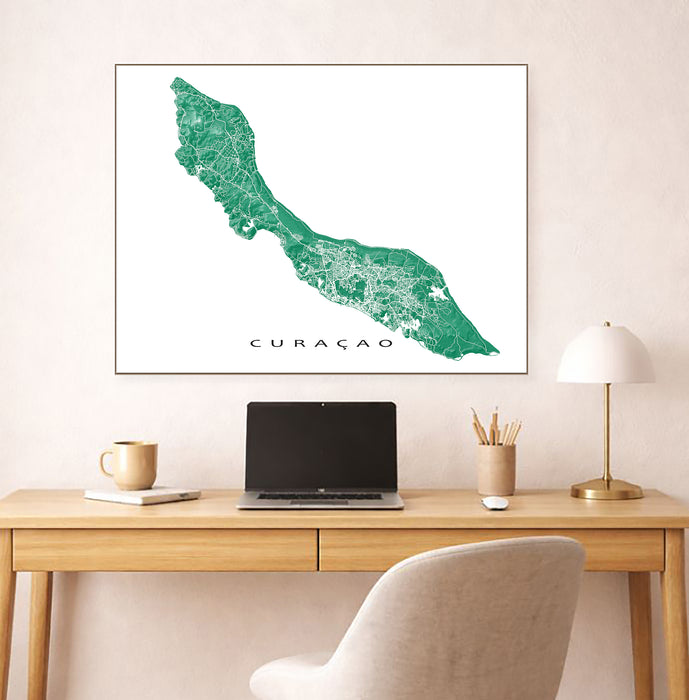 Framed map of Curacao on a wall above a desk with a laptop and lamp.