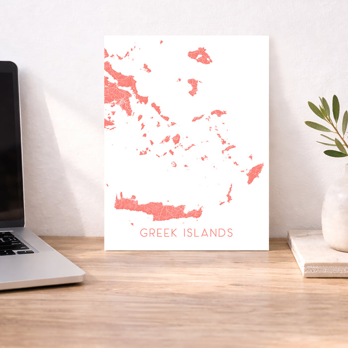 Map of Greek Islands on a white background displayed on a wooden desk with a laptop and plant.