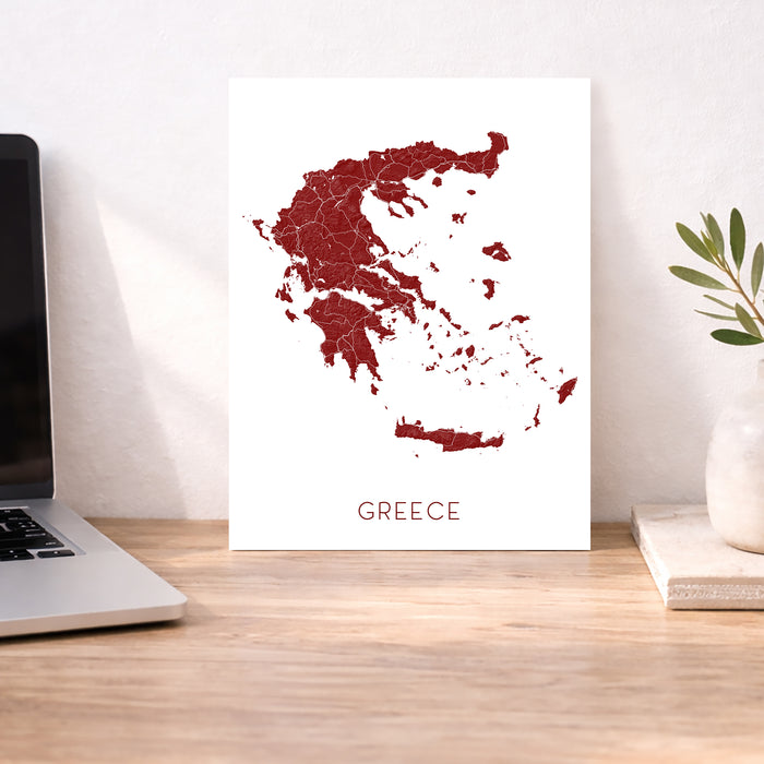 Map of Greece on a white background next to a laptop on a wooden desk.