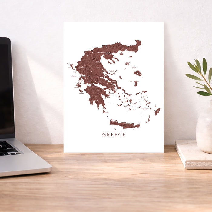 Map of Greece on a white background next to a laptop on a wooden desk.