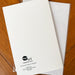 A white back side of a greeting card with the Maps As Art logo printed at the bottom, placed on top of a white envelope on a wooden surface.