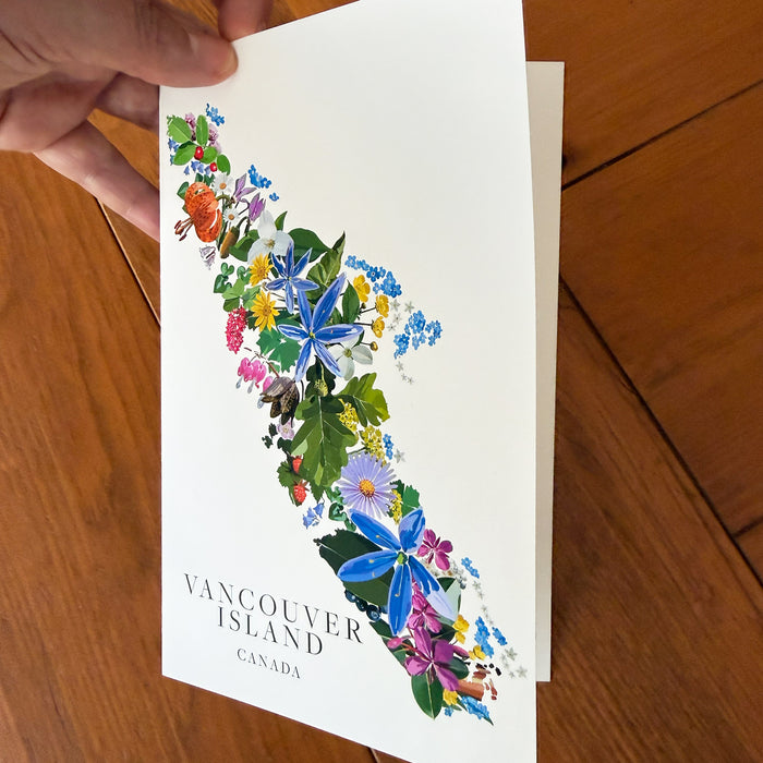 A hand holds a white greeting card featuring a colorful floral illustration of Vancouver Island, Canada, against a warm wooden tabletop background.